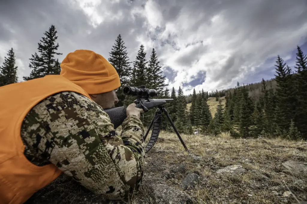 Atlas bipod deployed beneath a bolt-action rifle as a prone hunter aims across a pine-covered mountain valley.