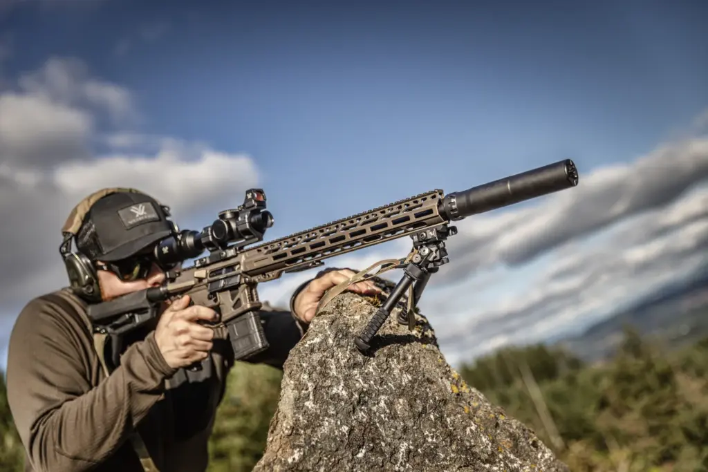 Shooter braced on a rock aiming a suppressed AR with the Atlas PSR BT46-LW17 bipod