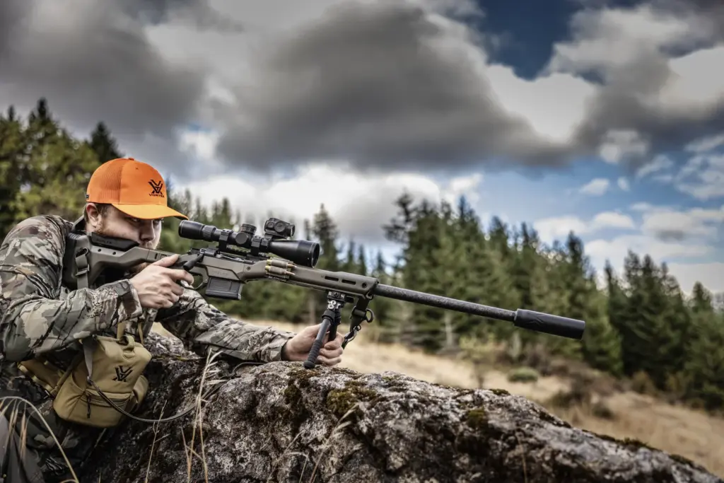 Hunter in blaze orange aiming a suppressed rifle from a rock outcrop on the Atlas PSR BT46-LW17 bipod