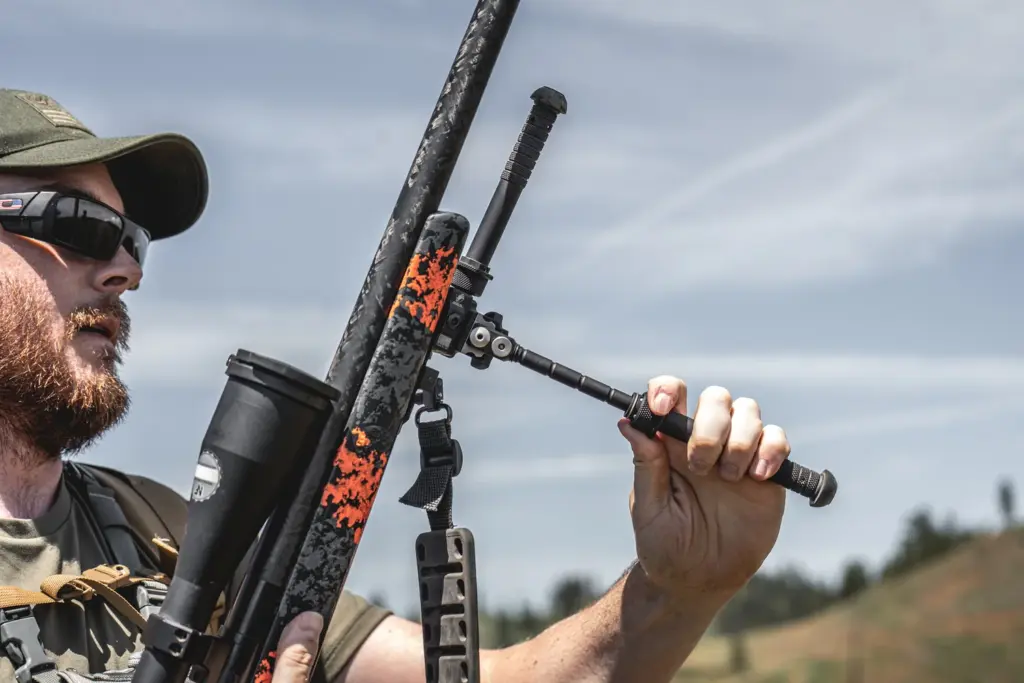 Shooter carrying an orange-camo rifle gripping the folded Atlas PSR BT46-LW17 bipod